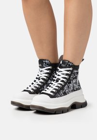 Sneakers alta nere con motivi grafici bianchi, lacci bianchi e una spessa suola bianca. Parte superiore in tela testurizzata e suola in gomma.