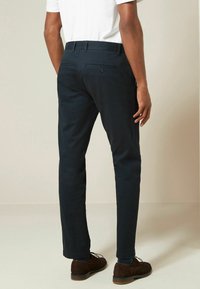 Marineblaues Baumwollhosen mit schmaler Passform, ausgestattet mit zwei Gesäßtaschen mit Knöpfen und einer glatten Textur. Kombiniert mit braunen Schuhen.