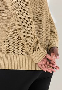 Maglione lavorato a maglia marrone chiaro con un design a rete aperta, polsini e orlo a coste, che mette in risalto una finitura testurizzata. Retro delle mani poggiate vicino alla vita.