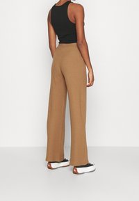 Pantalon large en maille côtelée marron, associé à des baskets noires et blanches, avec un débardeur noir ajusté. Fond blanc uni.