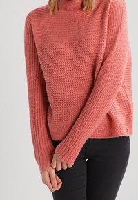 Pull tricoté en corail doux avec un col haut, une coupe ample et un motif texturé. Poignets et ourlet côtelés, fente latérale, et marquage minimaliste.