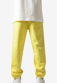 Pantaloni de trening galbeni cu manșete elastice, textură netedă și detalii pe cusăturile laterale. Asortați cu un tricou alb.