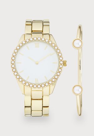 Montre dorée avec un cadran blanc, des chiffres romains et des accents en perles autour de la lunette. Inclut un bracelet doré assorti avec des détails en perles.
