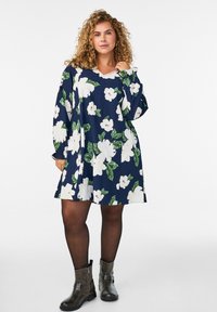 Blumenkleid mit navy Hintergrund, weißen Blumen und grünen Blättern. Lange Ärmel und lockere Passform, kombiniert mit dunklen Knöchelstiefeln.