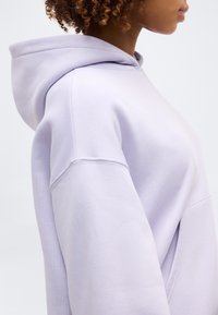 Γυναίκα με σγουρά μαλλιά που φοράει ένα ανοιχτό μωβ φαρδύ φούτερ με κουκούλα, εμφανιζόμενη πλάγια με την κουκούλα κατεβασμένη.