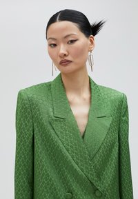 Blazer verde de doble botonadura en tejido texturizado, con cuello de muesca y detalles de botones, adornado con un sutil patrón en zigzag.