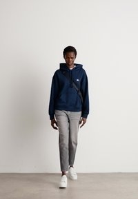 Mørkeblå hettegenser med lommer foran, brukt over en hvit skjorte, sammen med lysegrå jeans og hvite joggesko, stående på en jevn overflate.