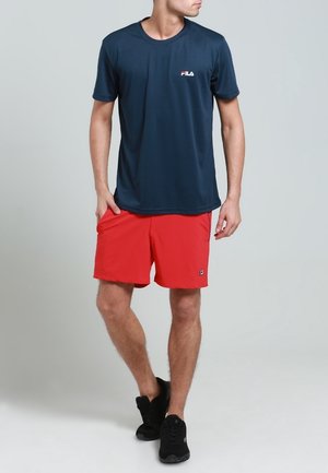Tmavě modré tričko odvádějící vlhkost s malým bílým a červeným logem, doplněné červenými šortkami. Celý vzhled dotváří černé sportovní boty.