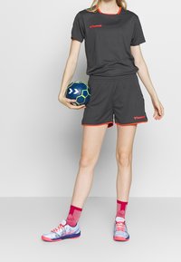 Uniforme deportivo gris con acentos rojos, que incluye una camiseta y pantalones cortos. El modelo sostiene un balón de mano azul y verde. Zapatillas deportivas azul y rosa.