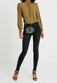 Blusa de cuello alto en verde oliva con mangas abullonadas, jeans ajustados negros y un bolso cruzado con cuentas y correa de cadena, combinado con tacones negros con tiras.