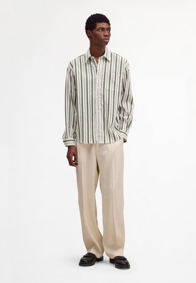Homme debout portant une chemise à rayures verticales vertes et blanches à boutons, un pantalon beige à jambes larges et des mocassins noirs, regardant vers la droite.