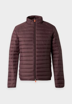 Homme portant une veste matelassée bordeaux, un pull bordeaux, un pantalon noir et des baskets blanches, debout avec une main dans la poche.