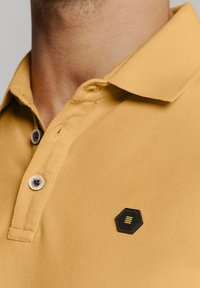 Geel polo shirt van een gestructureerde stof, met een zwart hexagonaal patch met gele accenten en twee knoopsluitingen.