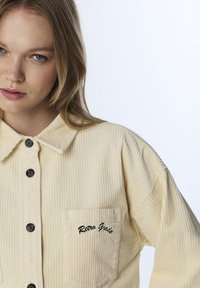 Corduroy shirt in pale yellow with a textured ribbed pattern, button closure, and left chest pocket featuring black embroidered text.
