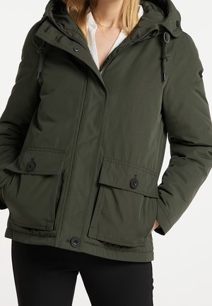Femme portant une veste d'hiver à capuche vert foncé avec de grandes poches avant boutonnées et une chemise blanche en dessous.
