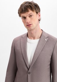 Brauner strukturierter Blazer in taillierter Form mit Reverskragen, einreihigem Knopfverschluss und einer Brusttasche, kombiniert mit einem weißen T-Shirt.