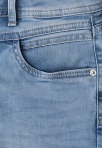 Poche de jean en denim bleu clair avec coutures visibles, un passant de ceinture au-dessus et un petit rivet métallique sur la couture latérale.