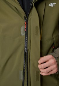 Personne portant une veste vert olive fermant une fermeture éclair noire avec une languette rouge, montrant des patchs Velcro et une partie d'une main.