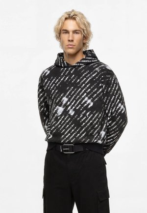 Giovane uomo con capelli biondi mossi che indossa una felpa nera con testo diagonale bianco e pantaloni neri con cintura, in piedi davanti a uno sfondo bianco.