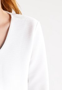 Top de tela blanca con textura acanalada y escote en V. Presenta un brillo sutil y mangas estructuradas. Detalle texturizado visible a lo largo de la tela.