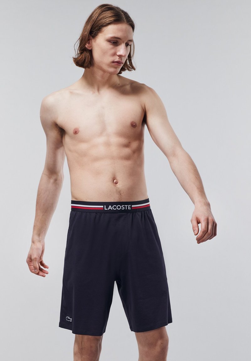 Pantaloni corti blu navy con vita elasticizzata, dotati di logo Lacoste e strisce rosse e bianche. Texture liscia e design lunghezza ginocchio.