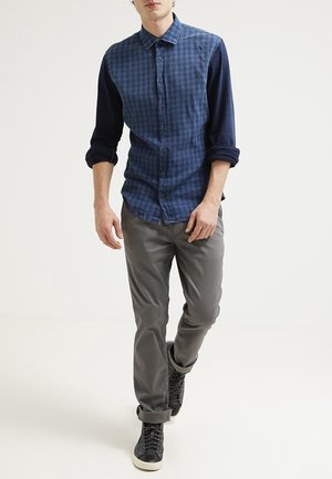 Mann, der ein blau kariertes Hemd mit Knöpfen, graue Hosen und schwarze hochgeschnittene Turnschuhe trägt und auf weißem Hintergrund geht.