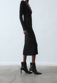 Robe noire à manches longues côtelées avec un décolleté en V profond, silhouette ajustée et longueur mi-mollet, associée à des bottines noires à talons.