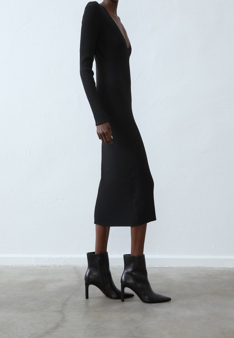 Robe noire à manches longues côtelées avec un décolleté en V profond, silhouette ajustée et longueur mi-mollet, associée à des bottines noires à talons.