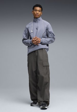 Jonge man die staat met gevouwen handen, gekleed in een grijze zip-up sweatvest, donkere cargobroek, zwarte sneakers en witte oordopjes die om zijn nek hangen.