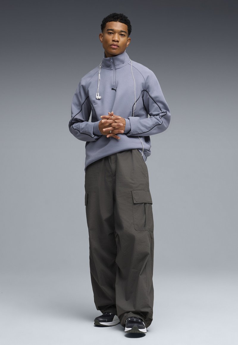 Jeune homme debout, les mains jointes, portant un sweat-shirt gris à fermeture éclair, un pantalon cargo foncé, des baskets noires et des écouteurs blancs autour du cou.