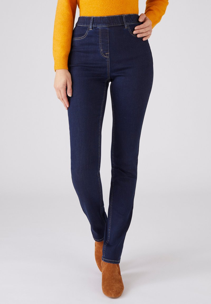 Jeans de denim de color azul oscuro con una cintura alta, que presentan detalles de costura visibles y un corte ajustado. Llevados con botines de ante marrón.