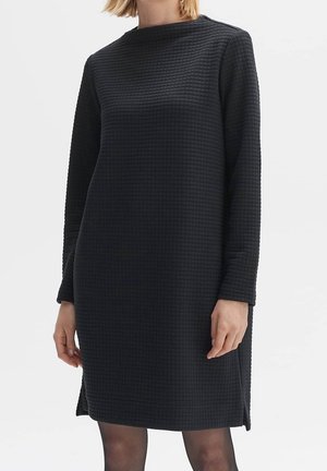Mujer con un vestido negro texturizado de manga larga hasta la rodilla, con corte recto y discretas aberturas laterales, de pie frente a un fondo liso.