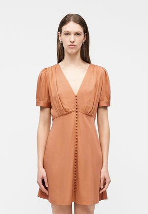 Femme aux cheveux bruns lisses portant une robe orange-brun à manches courtes, boutonnée à l'avant, avec un décolleté en V profond, debout devant un fond uni.