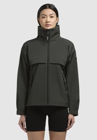 Giacca impermeabile verde scuro con colletto alto, chiusura frontale con zip e grande tasca sul petto. Include tasche laterali e una vestibilità rilassata.