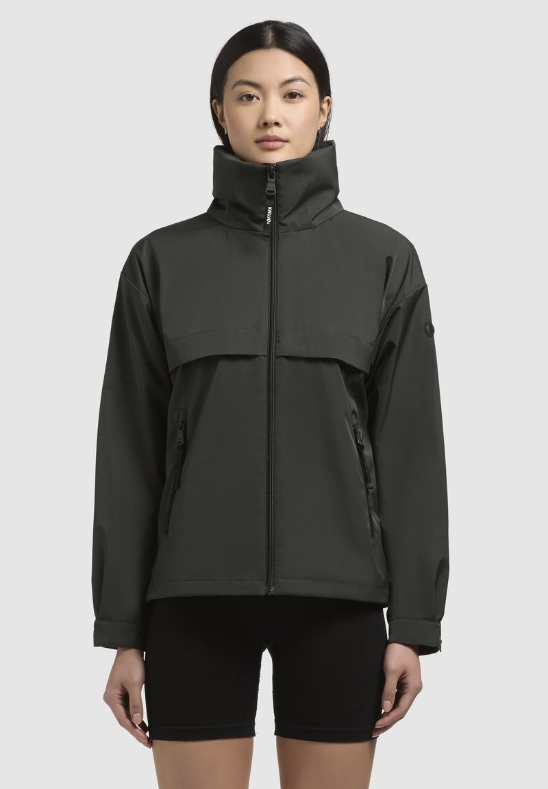 Giacca impermeabile verde scuro con colletto alto, chiusura frontale con zip e grande tasca sul petto. Include tasche laterali e una vestibilità rilassata.