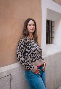 Strickpullover mit beigem Grund und schwarzem Leopardenmuster, gerippte Bündchen und Saum in Rot. Kombiniert mit blauen Jeans.