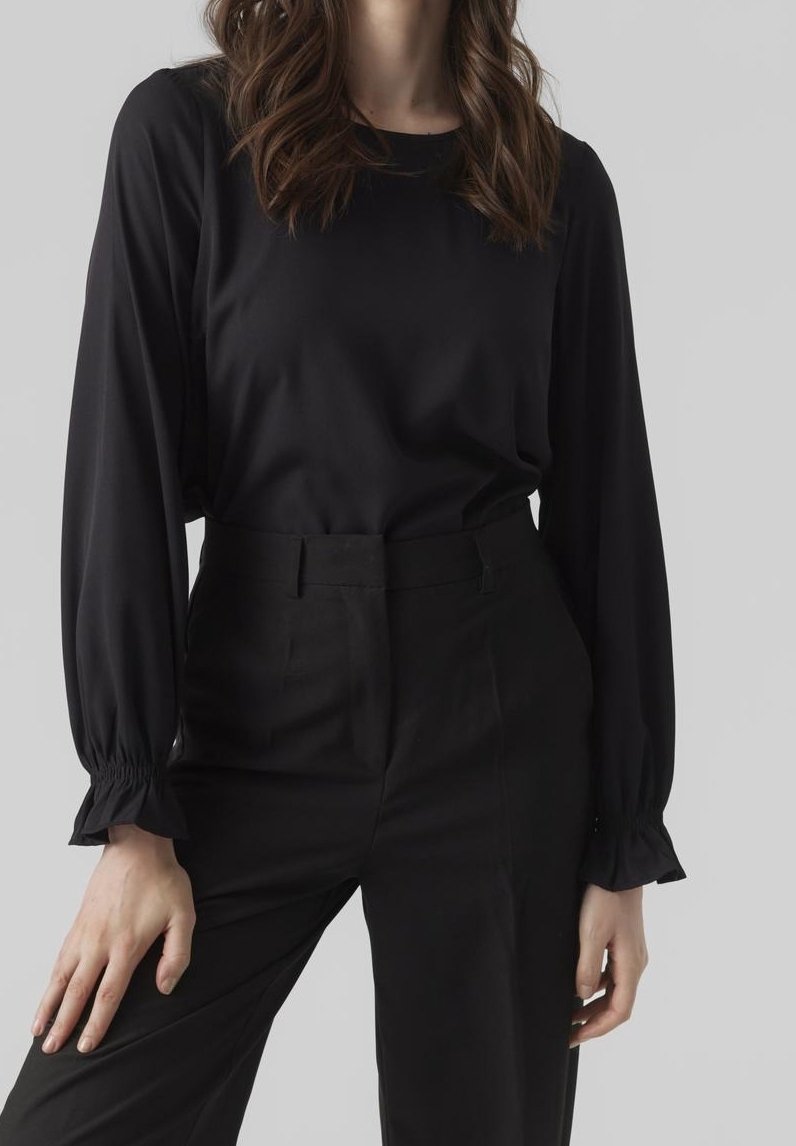 Femme portant un chemisier noir à manches longues avec poignets froncés et un pantalon noir taille haute sur un fond gris uni.