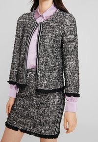 Svart och vit texturerad tweedjacka med fransar nertill, parat med en lila knapp-up skjorta med krage och långa ärmar.