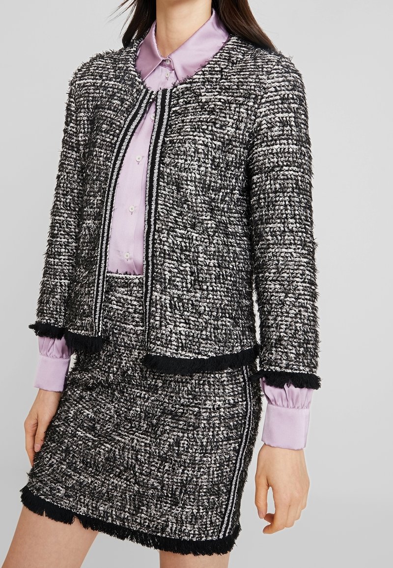 Svart och vit texturerad tweedjacka med fransar nertill, parat med en lila knapp-up skjorta med krage och långa ärmar.