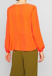 Top peplum di un brillante arancione con maniche lunghe a sbuffo, texture liscia e polsini elasticizzati. Abbinato a pantaloni a gamba larga di un verde oliva.