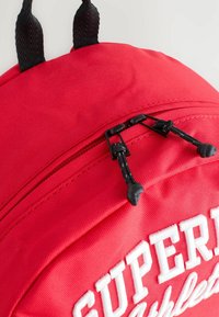 Röd tygryggsäck med en texturerad yta, dubbla svarta dragkedjehandtag och vit broderad text "SUPERB Athlete" på framsidan.