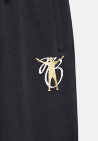 Czarne dresy z fakturowanej tkaniny. Złota i biała grafika przedstawiająca postać z wyciągniętymi ramionami znajduje się na lewym nogawce.