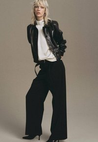 Black leather bomber jacket over a white turtleneck, paired with high-waisted black wide-leg trousers and pointed black heels.
