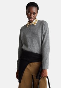 Šedý vlněný svetr s žebrovanými manžetami, vrstvený na kostkované žluté a bílé košili, doplněný o hnědé kalhoty. Černý svetr uvázaný kolem pasu.