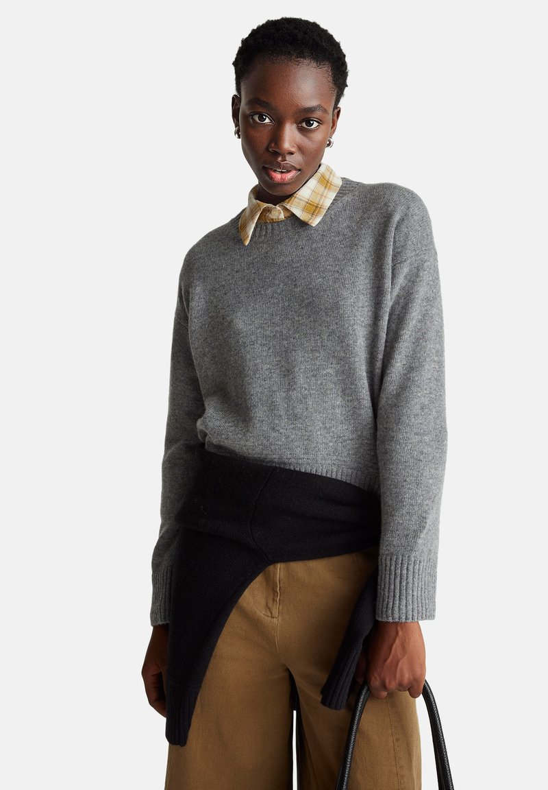 Šedý vlněný svetr s žebrovanými manžetami, vrstvený na kostkované žluté a bílé košili, doplněný o hnědé kalhoty. Černý svetr uvázaný kolem pasu.