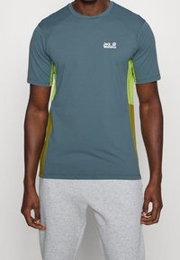 Tričků s krátkým rukávem v barvě teal s limetkově zelenými bočními panely. Obsahuje kulatý výstřih a malé bílé logo na hrudi. Hladká textura.