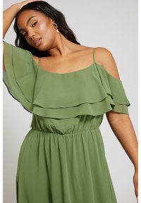 Grön, lager på lager chiffongklänning med off-the-shoulder design, elastisk midja och mjuk textur; har justerbara axelband och volanger.