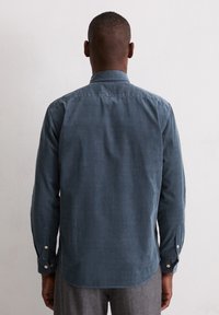 Mann mit langärmliger, dunkelblauer Druckknopfbüroschürze und grauen Hosen, der vor einer weißen strukturierten Wand steht und mit dem Rücken zur Kamera blickt.