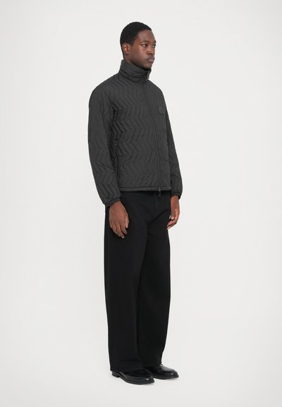 Man standing wearing a black quilted zip-up jacket, wide-leg black pants, and black leather shoes against a plain white background.