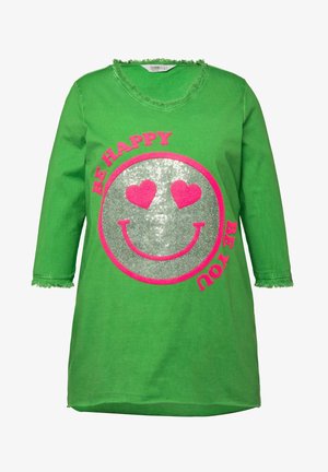 Túnica de algodón verde con un escote con flecos. Presenta una gran cara sonriente plateada y corazones rosas, acompañada del texto "SE FELIZ".